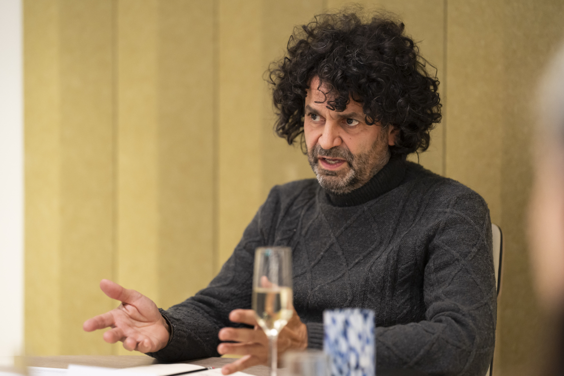 Senior designer engaged in discussion about technology's role in sustainable design, with glass and notebook in front.