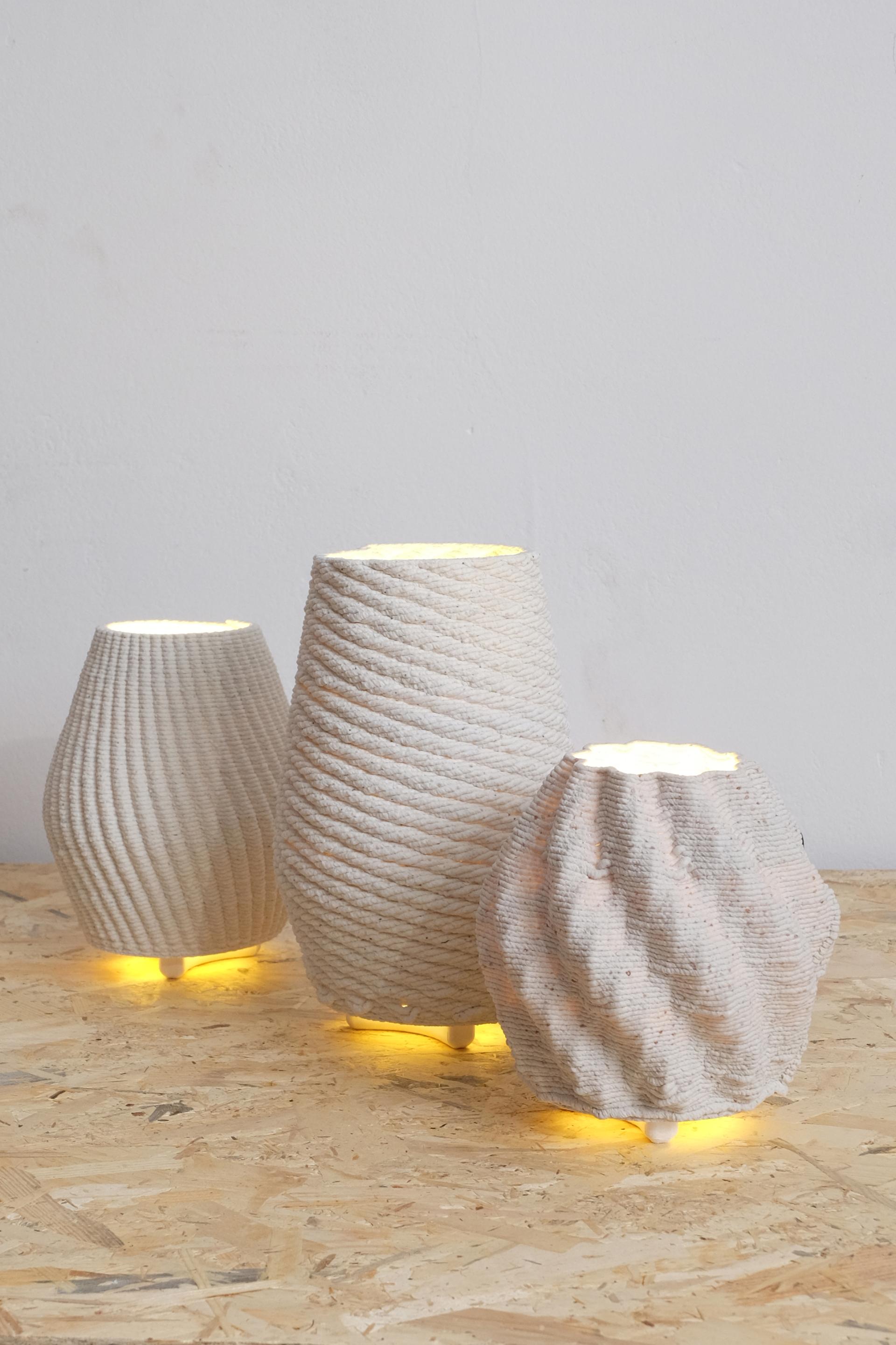 Three unique 3D printed lamps made from recycled ceramics, featuring intricate textures and warm glowing bases.