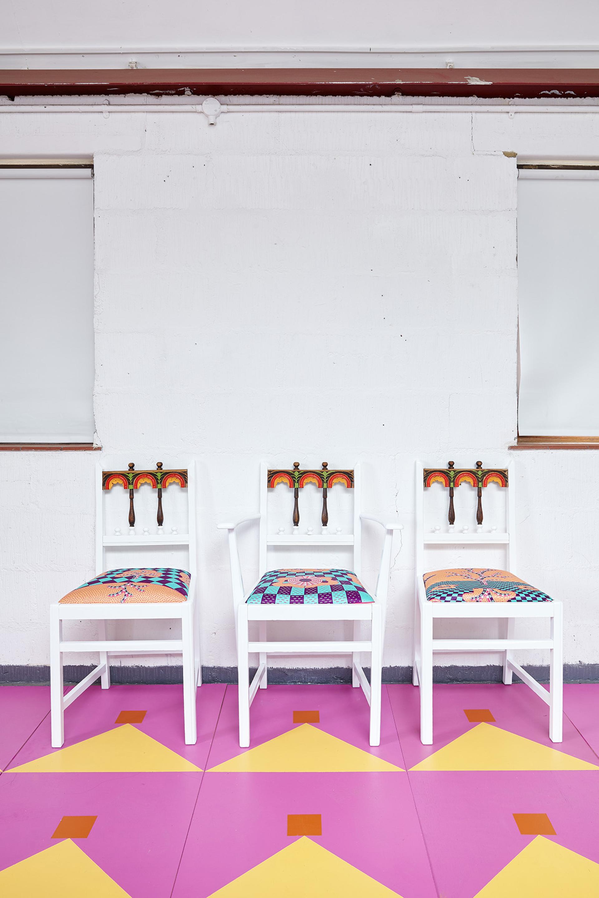 Colorful, upcycled chairs with intricate patterns, set against a minimalist white wall and vibrant pink floor design.
