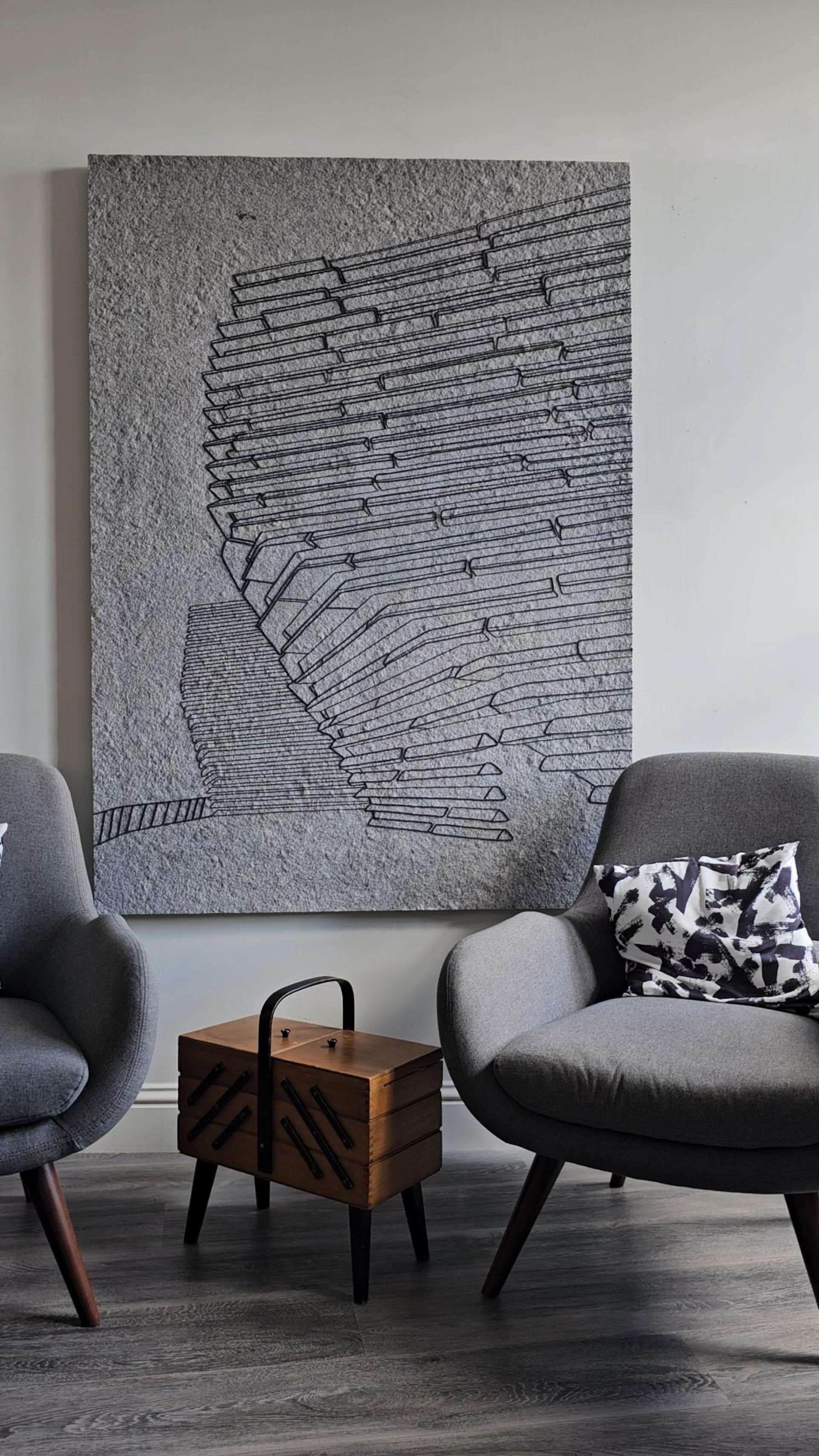 Textured textile artwork by Sandra Junele displayed on a wall, surrounded by modern armchairs and a wooden side table.