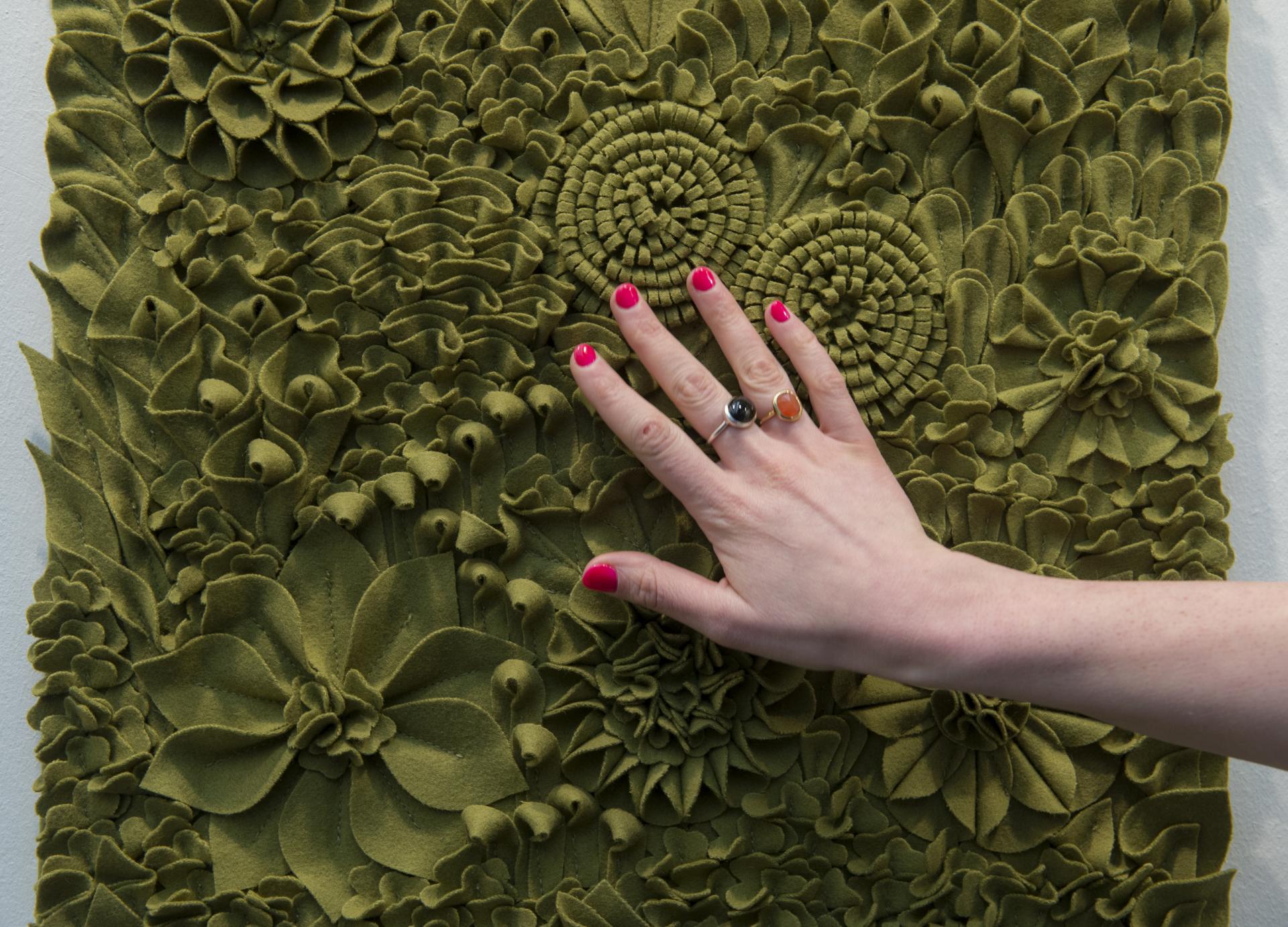A hand with bold pink nails touches a textured green wall covering featuring intricate floral designs at the Surface Design Show.