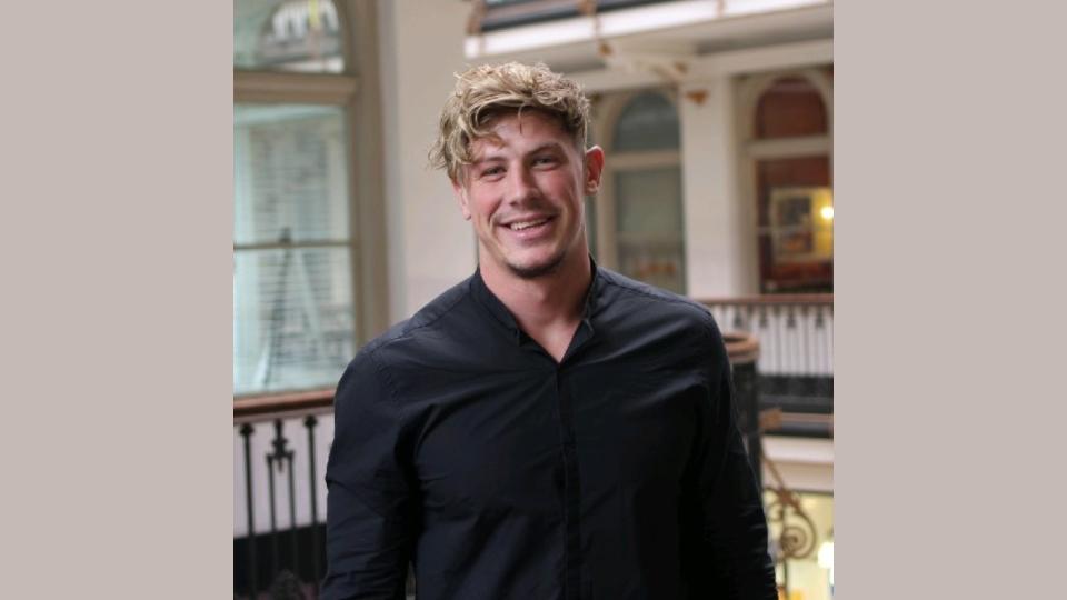 Tom Ailes from The Furniture Practice smiles confidently in a modern interior, reflecting innovative furniture design concepts.