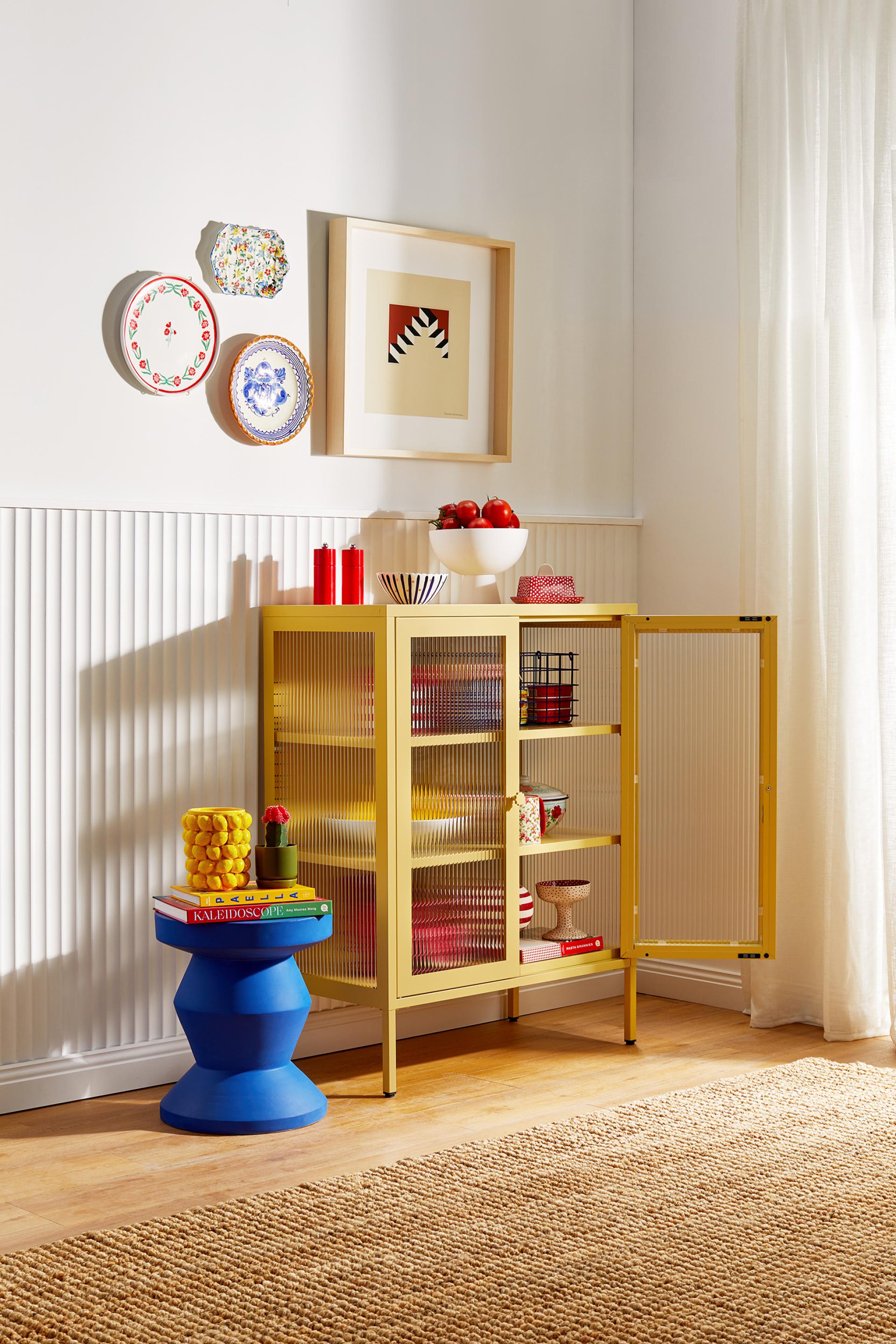 Vibrant yellow cabinet from Mustard Made's Glass Collection, showcasing playful design with colorful decor and plates on the wall.