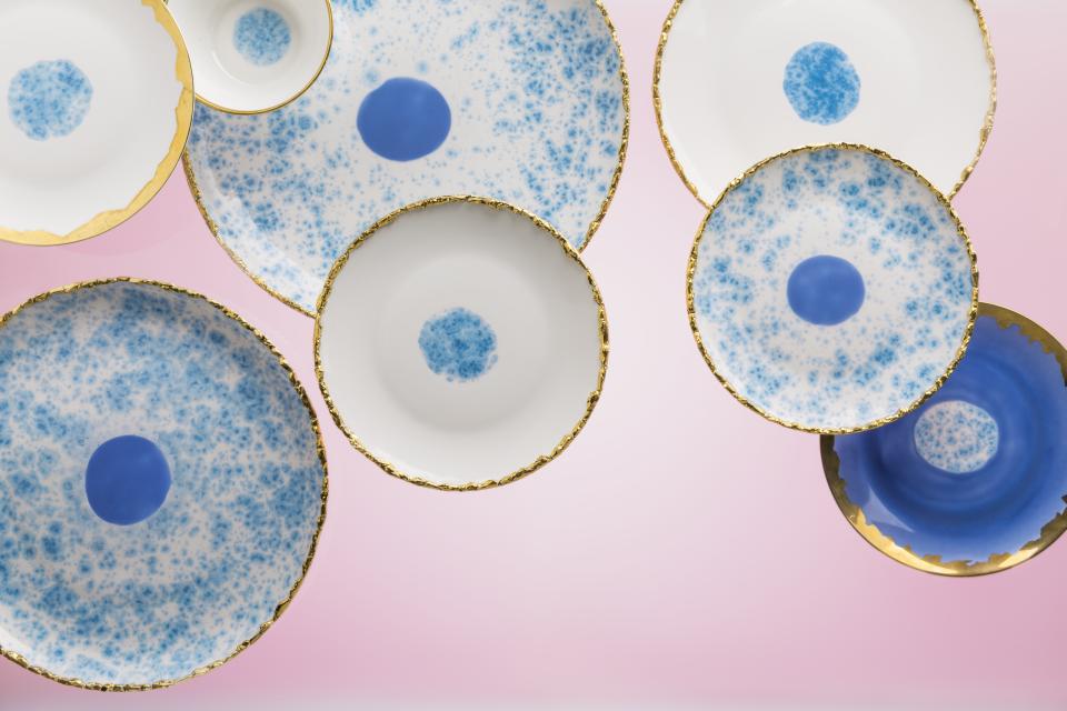 Hand-decorated ceramic and porcelain tableware by Coralla Maiuri featuring blue patterns and gold accents on a pink background.
