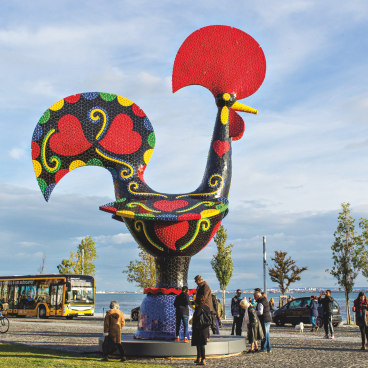 Colorful, oversized rooster sculpture by Joana Vasconcelos at a waterfront location, with visitors admiring the artwork.