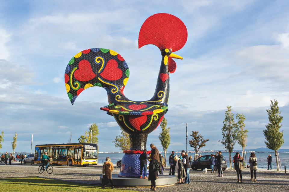 Colorful, oversized rooster sculpture by Joana Vasconcelos at a waterfront location, with visitors admiring the artwork.