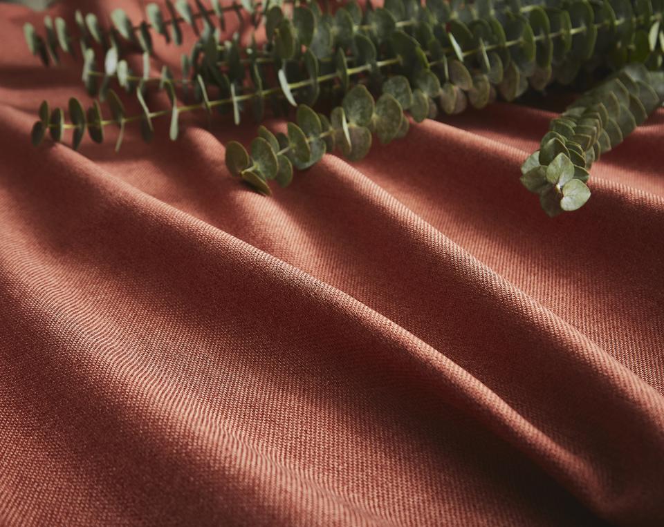 Rich terracotta fabric from the Tundra collection by ILIV, accented with eucalyptus leaves, showcasing its texture and vibrant color.