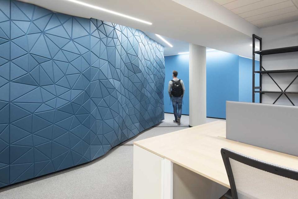 Modern office interior featuring a textured blue wall and a person walking past a desk area.