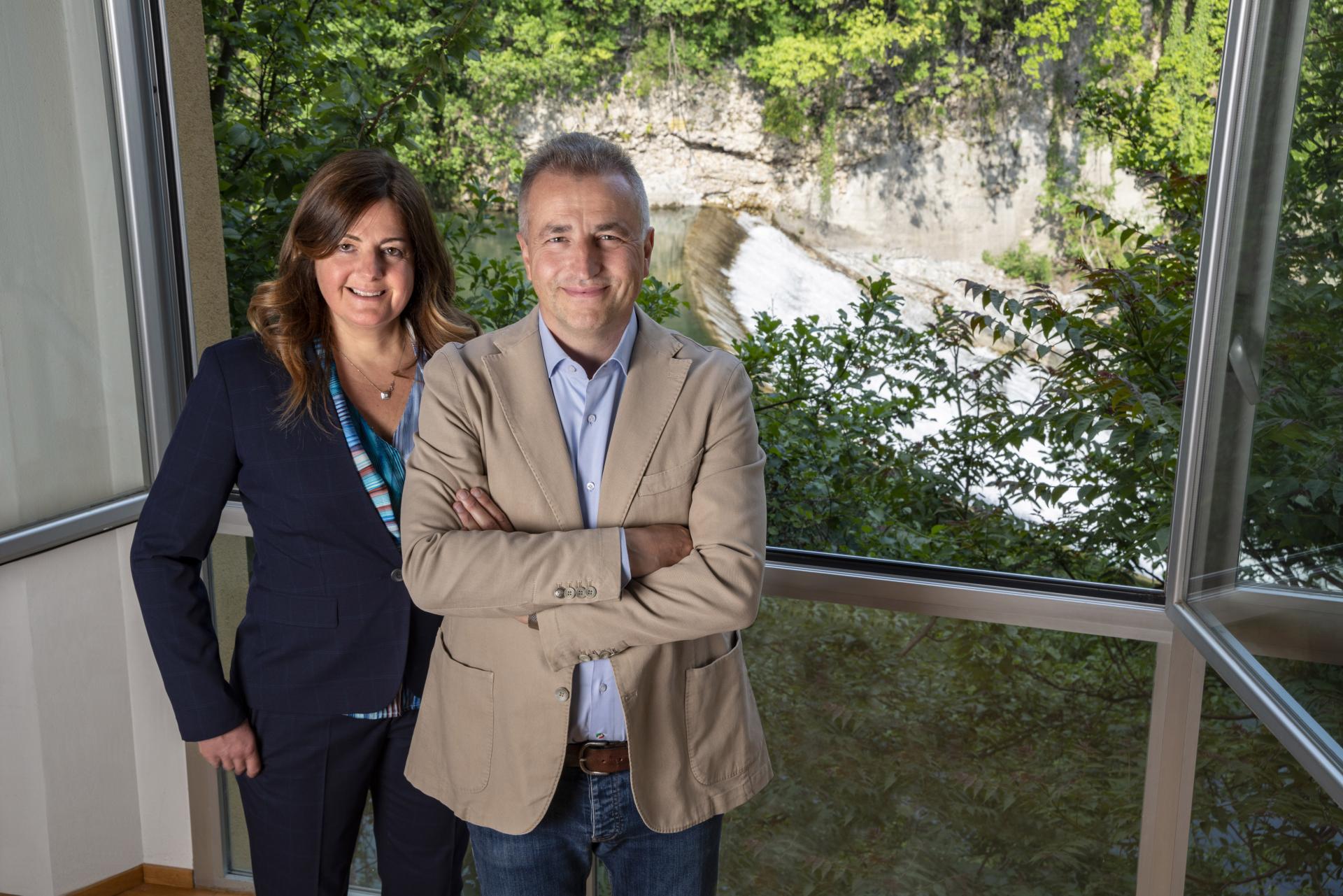 Founders of VIGANÒ & C. posing by a window overlooking a scenic waterfall, representing the brand's evolution and legacy.