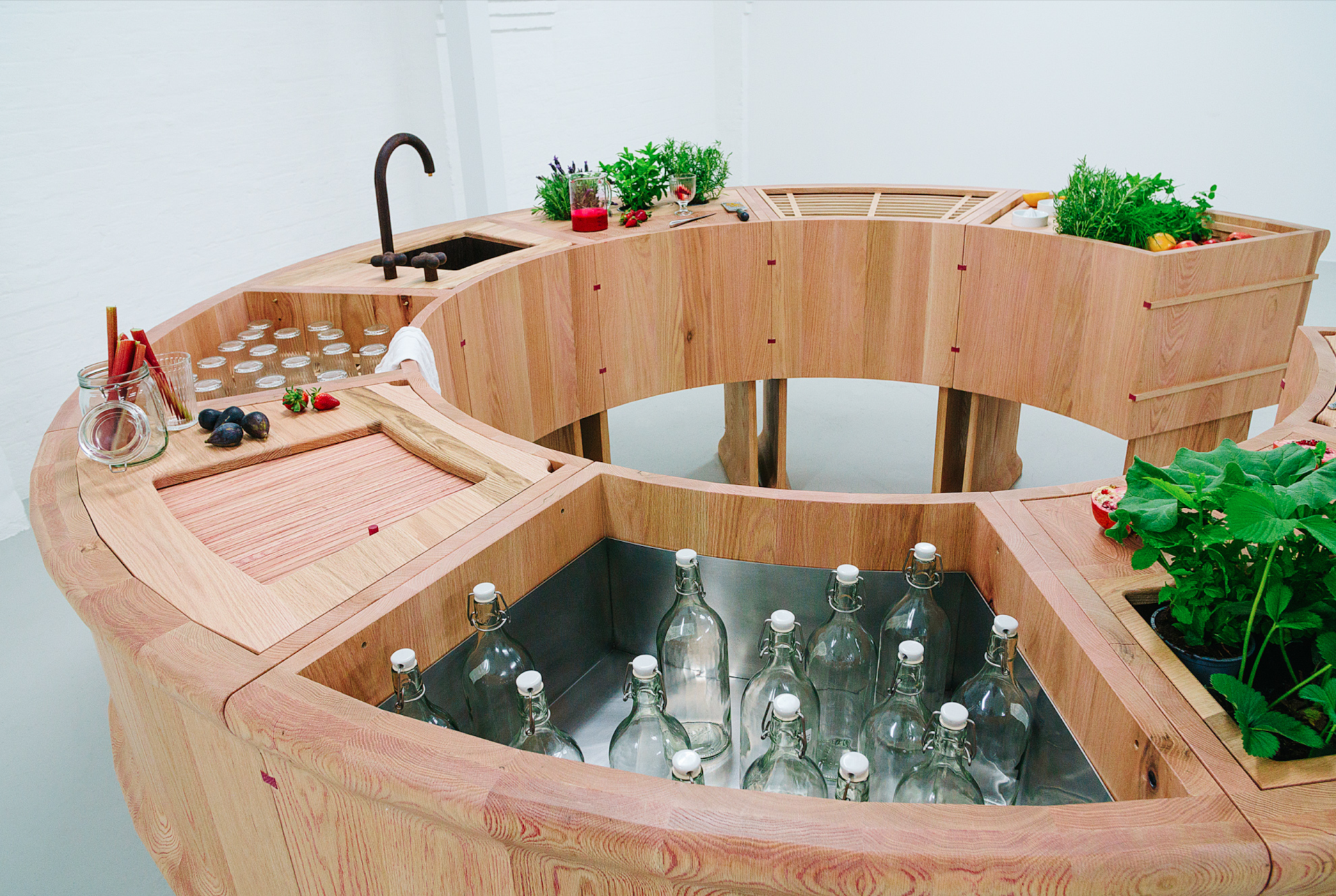 A round, red oak bar design featuring a sink, bottles, fresh herbs, and fruits, showcased at Milan Design Week.