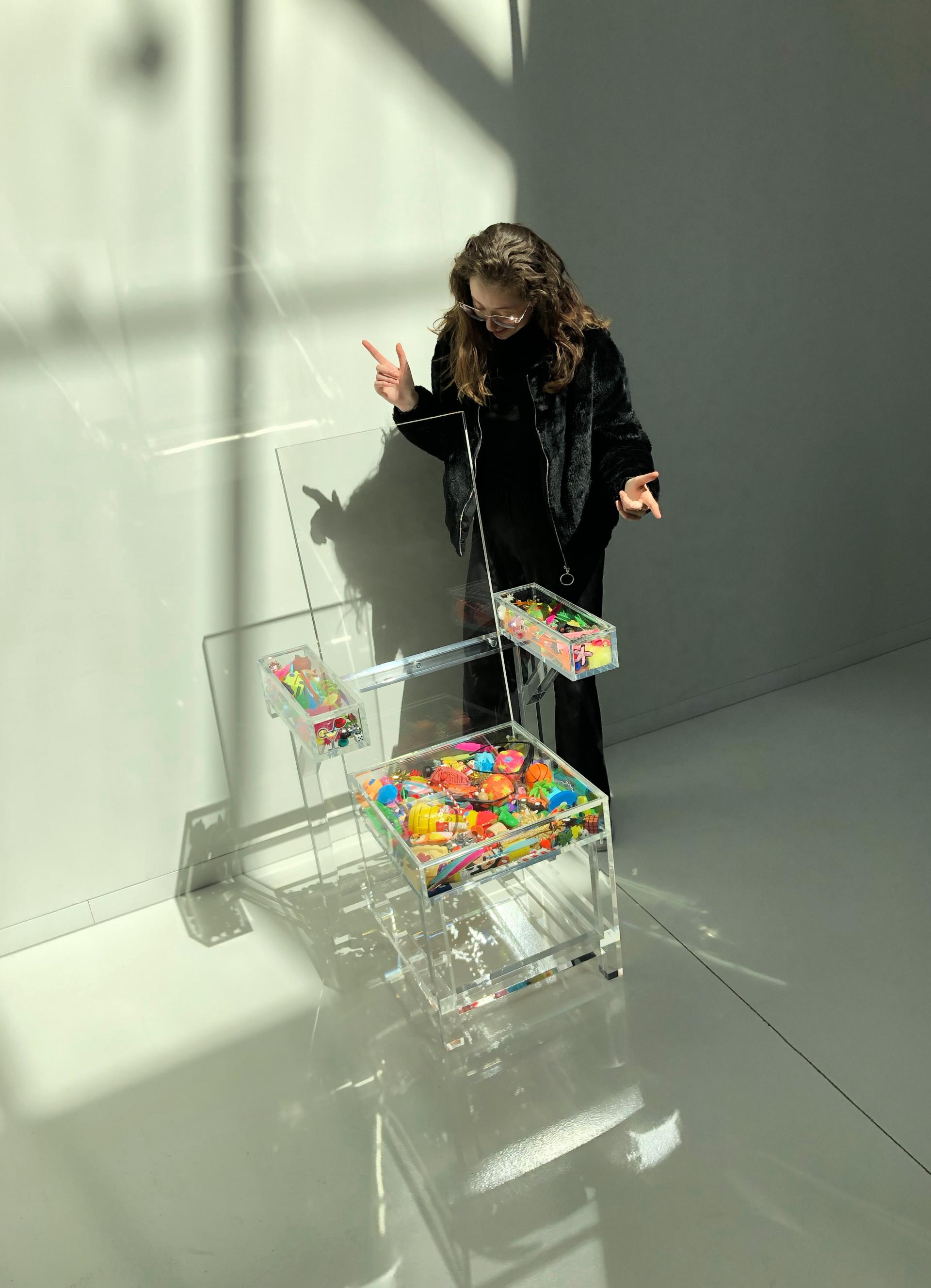 A female designer interacts with a colorful, transparent chair filled with playful toys, showcasing creativity and design innovation.