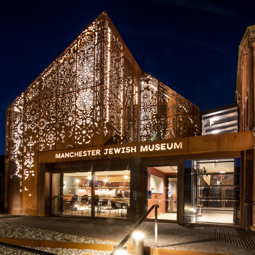 Manchester Jewish Museum features a contemporary corten steel façade illuminated at night, showcasing intricate design against a historic building.