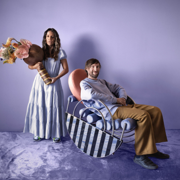 A woman in a flowing dress holds a large vase of flowers beside a man sitting in a unique modern chair.
