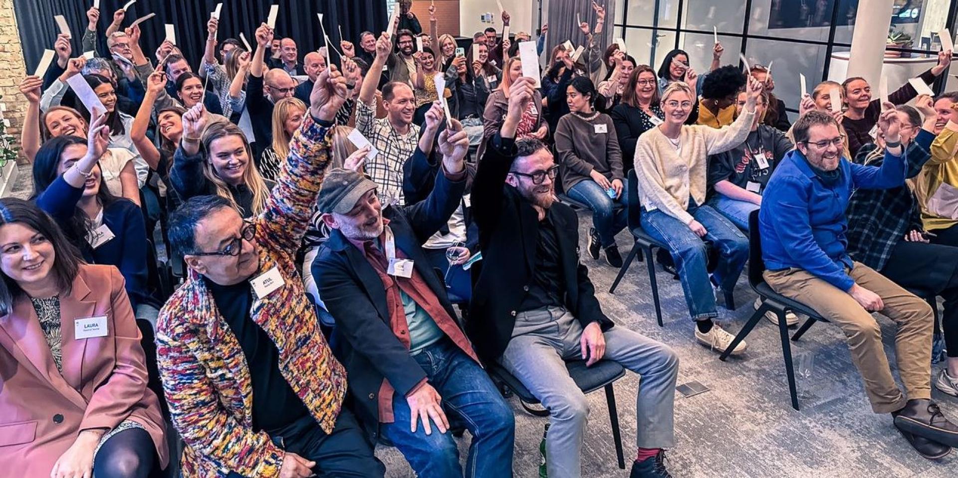 Attendees at a design event raise cards in support of EDI initiatives, reflecting engagement and collaboration in innovation.