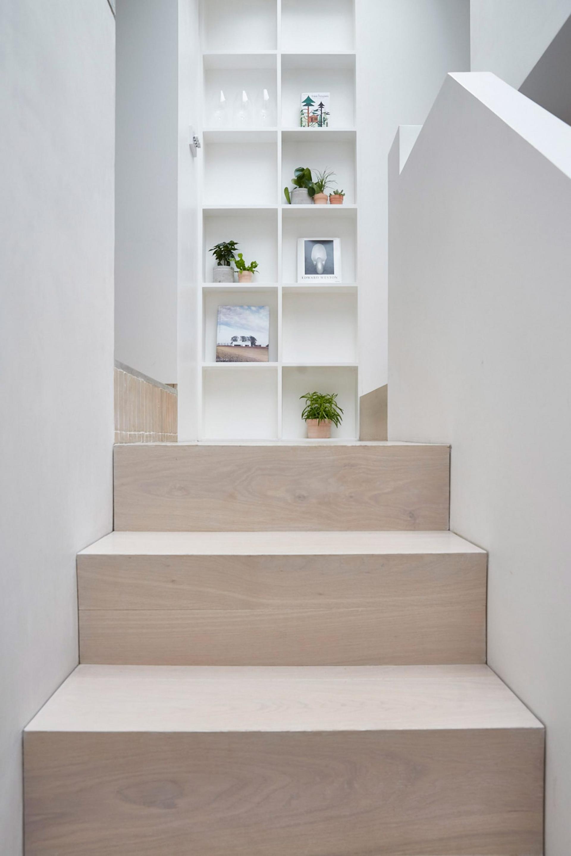 Light wooden stairs lead to a minimalist space featuring a white shelving unit decorated with plants and books.