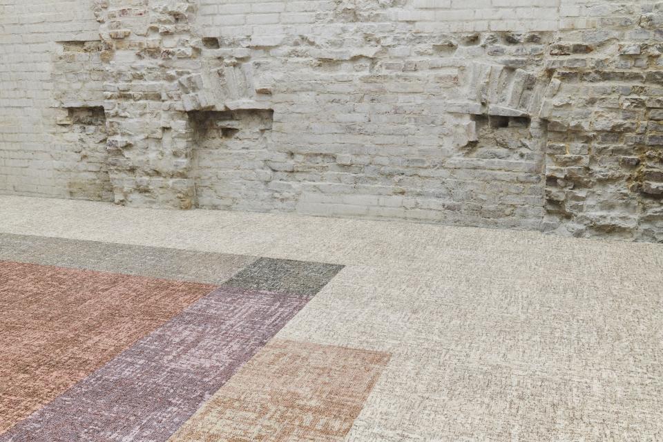 Modern carpet tiles in earthy tones laid out on a textured floor against a rustic brick wall, promoting wellbeing.
