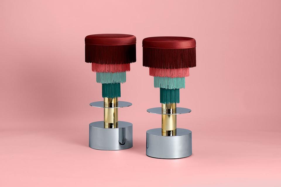 Colorful stools designed by Masquespacio, featuring layered fringe and metallic bases, set against a pink backdrop.