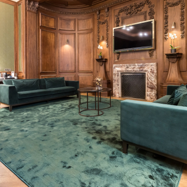 Luxurious green carpet covers the floor, complementing elegant wooden walls and plush velvet sofas in a premium hotel setting.
