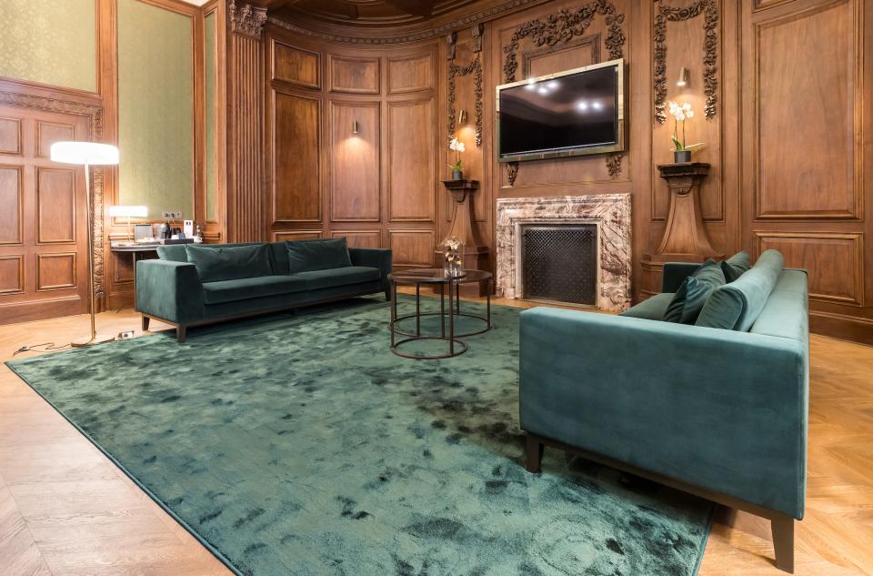 Luxurious green carpet covers the floor, complementing elegant wooden walls and plush velvet sofas in a premium hotel setting.