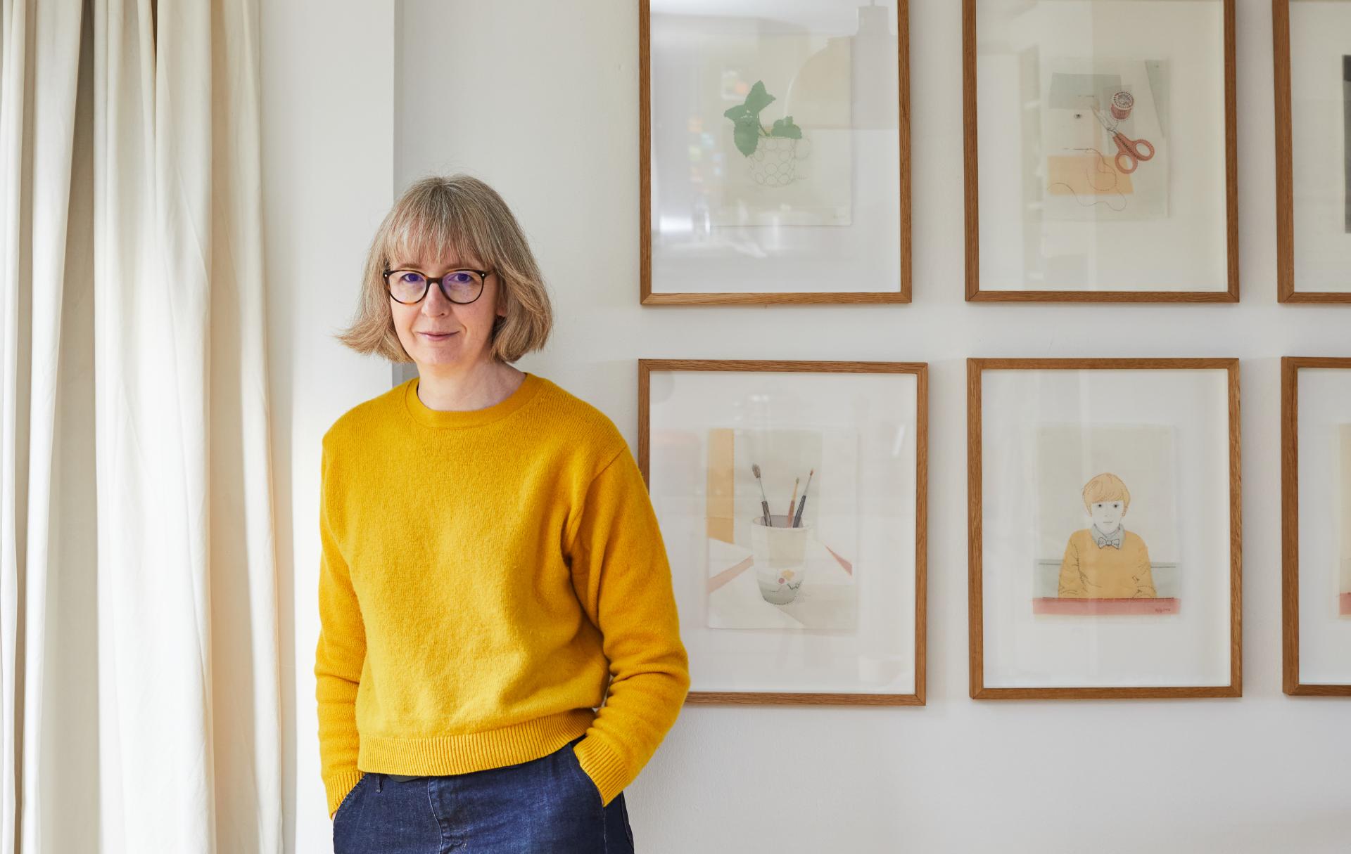 A designer-maker stands in a bright space, surrounded by framed artwork showcasing creativity and craft in design.