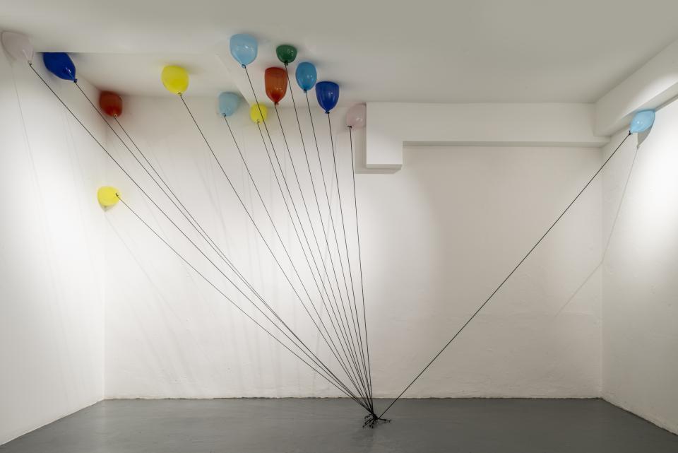 An installation featuring colorful glass balloons tethered by black strings in a minimalist gallery space.