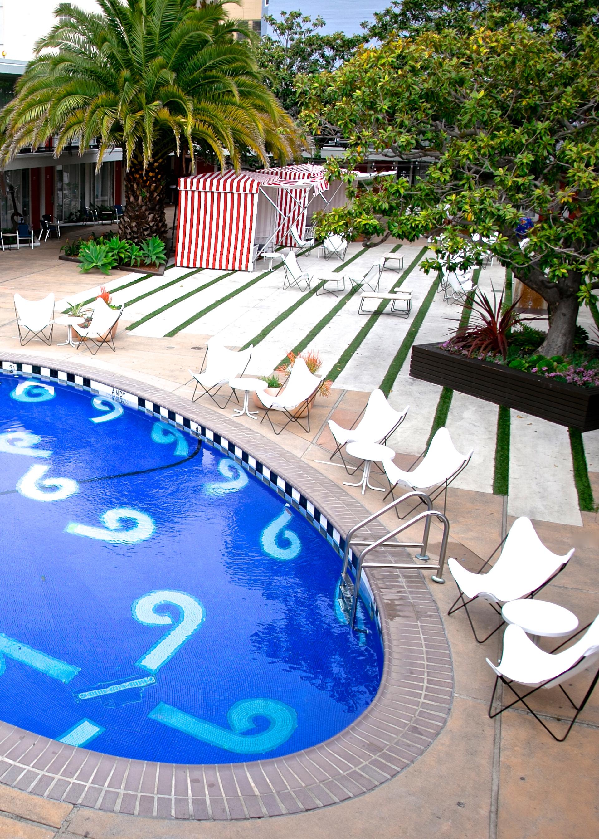 Vibrant blue pool with decorative patterns, surrounded by white chairs, palm trees, and striped cabanas at Phoenix Hotel.