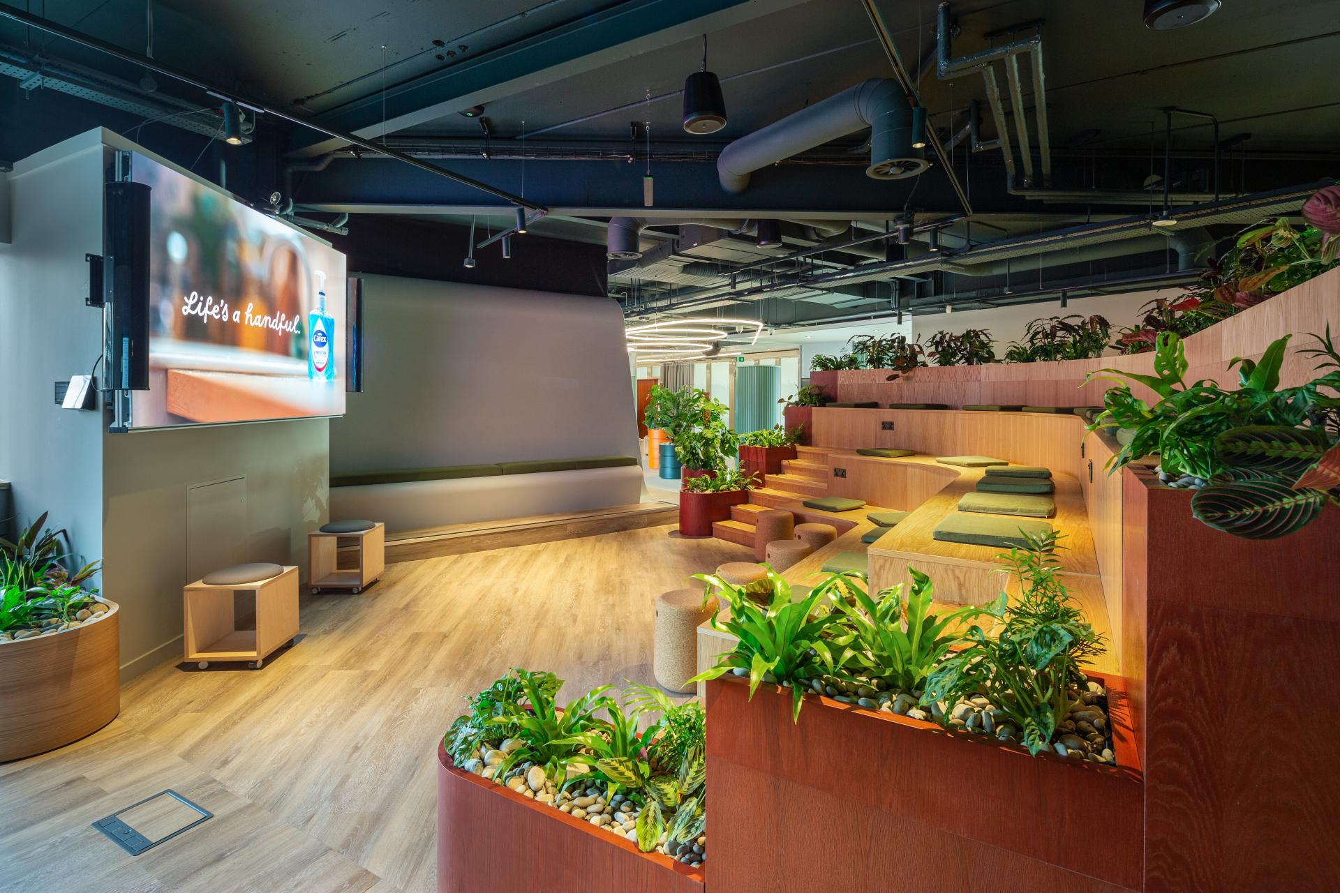 Modern interior design space featuring lush greenery, comfortable seating, and a large screen displaying an uplifting message.