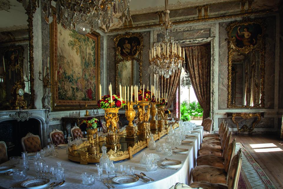 Elegant dining room featuring a grand table, intricate chandeliers, ornate decorations, and lavish floral centerpieces in a historic British interior.