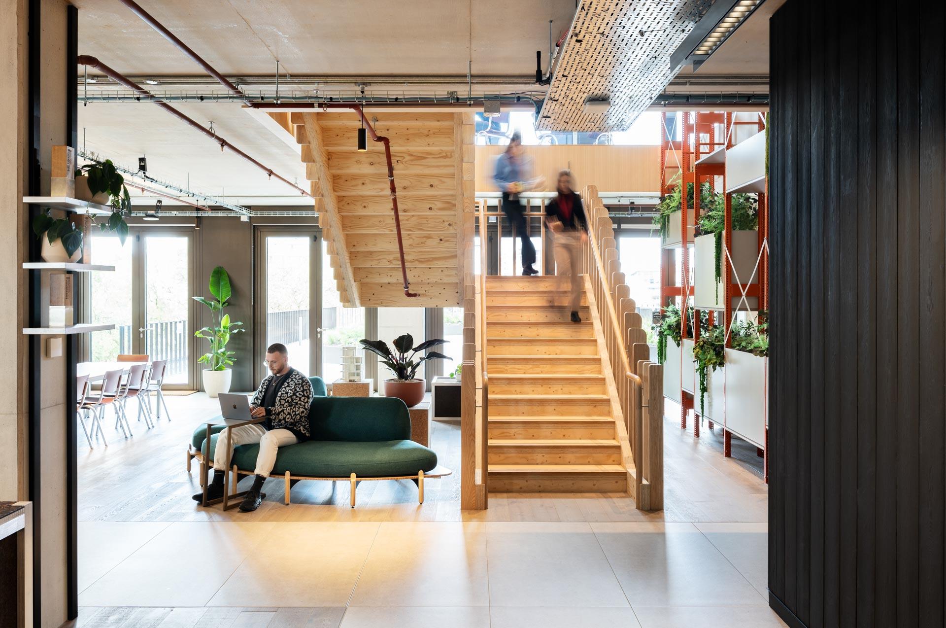 Modern workplace interior featuring a green sofa, wooden staircase, and plants, reflecting design principles of nature and neurodiversity.
