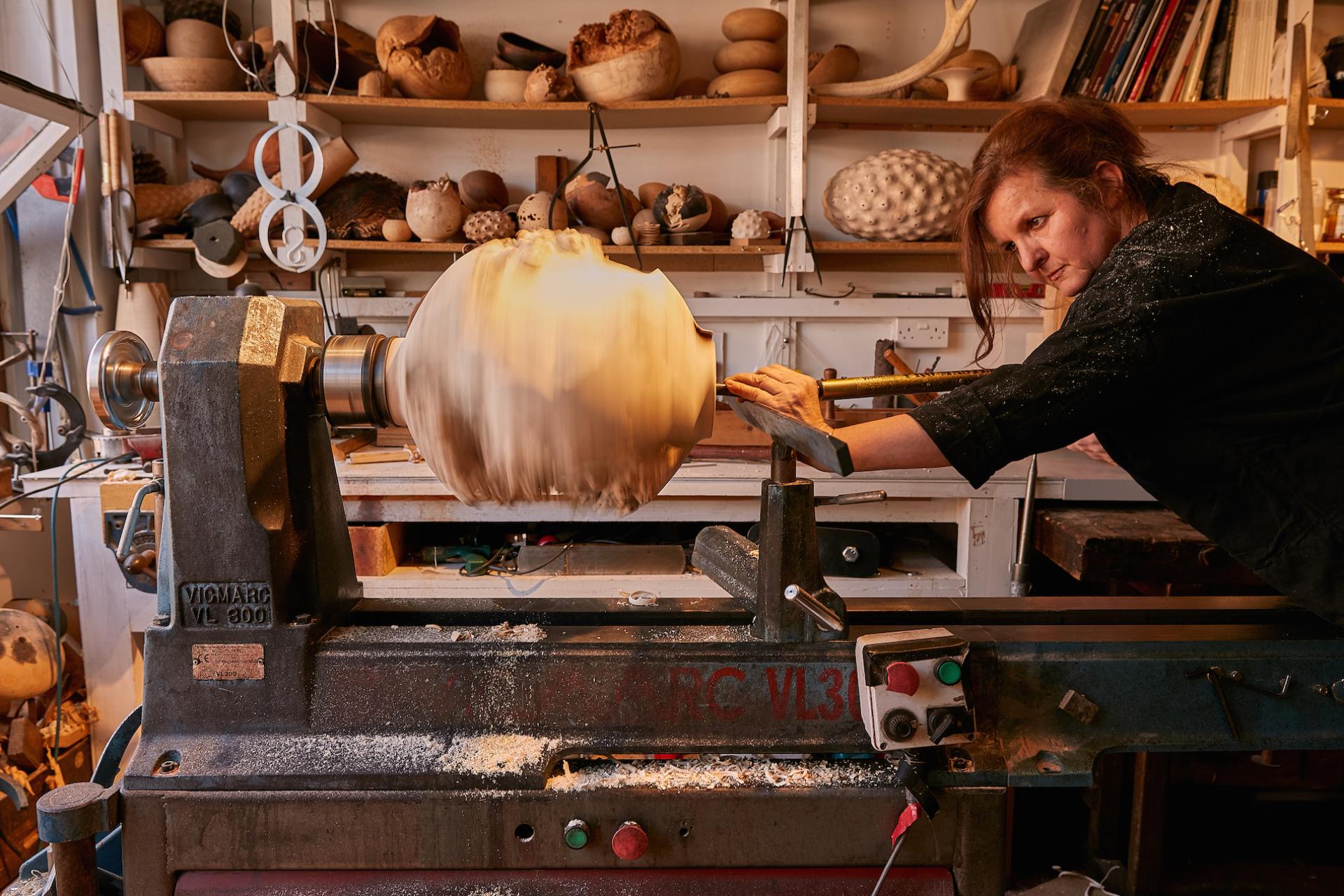 Craftsperson using a lathe to shape wood, surrounded by artistic creations in a busy workshop during Cockpit Arts Summer Festival.