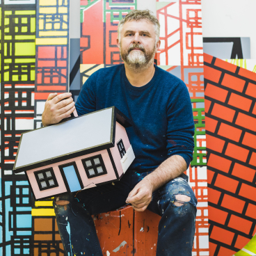 Artist Richard Woods holds a colorful model house, surrounded by vibrant painted backdrops, showcasing his inventive work with raw materials.