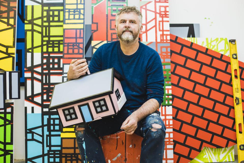 Artist Richard Woods holds a colorful model house, surrounded by vibrant painted backdrops, showcasing his inventive work with raw materials.