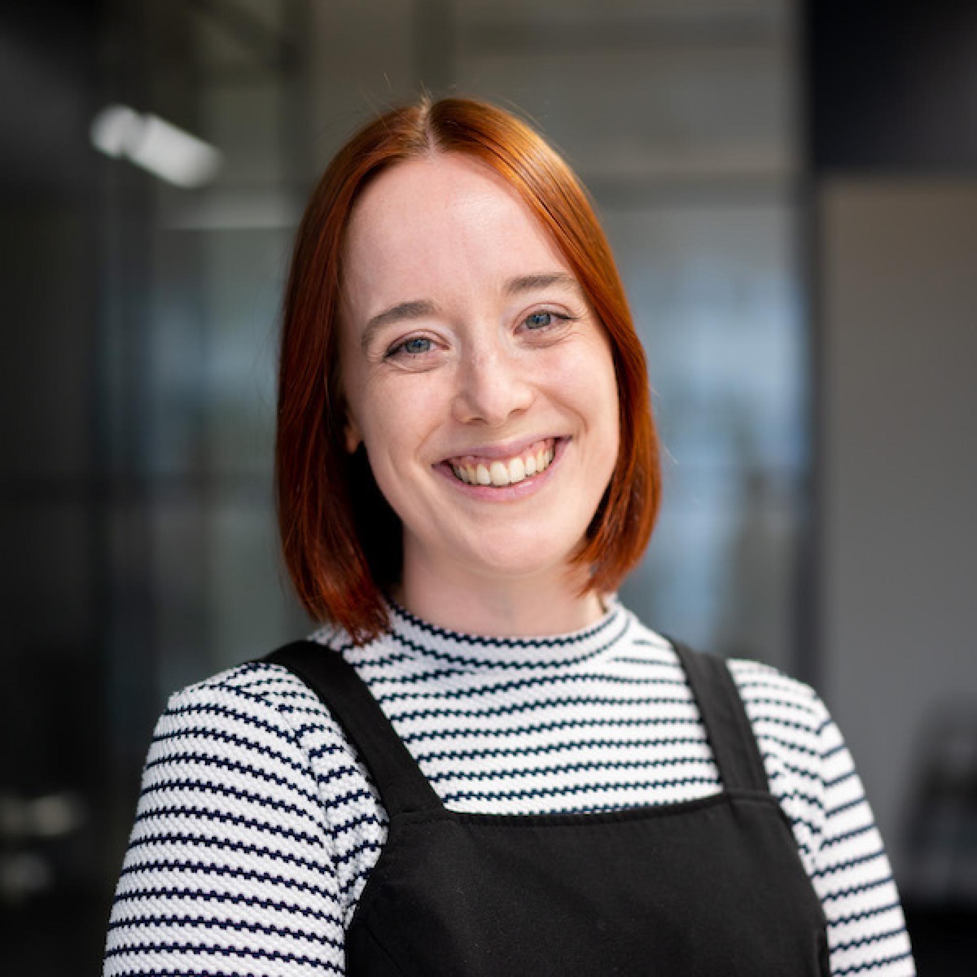 Smiling woman with red hair wearing a striped shirt and black overall, representing a modern workplace environment.