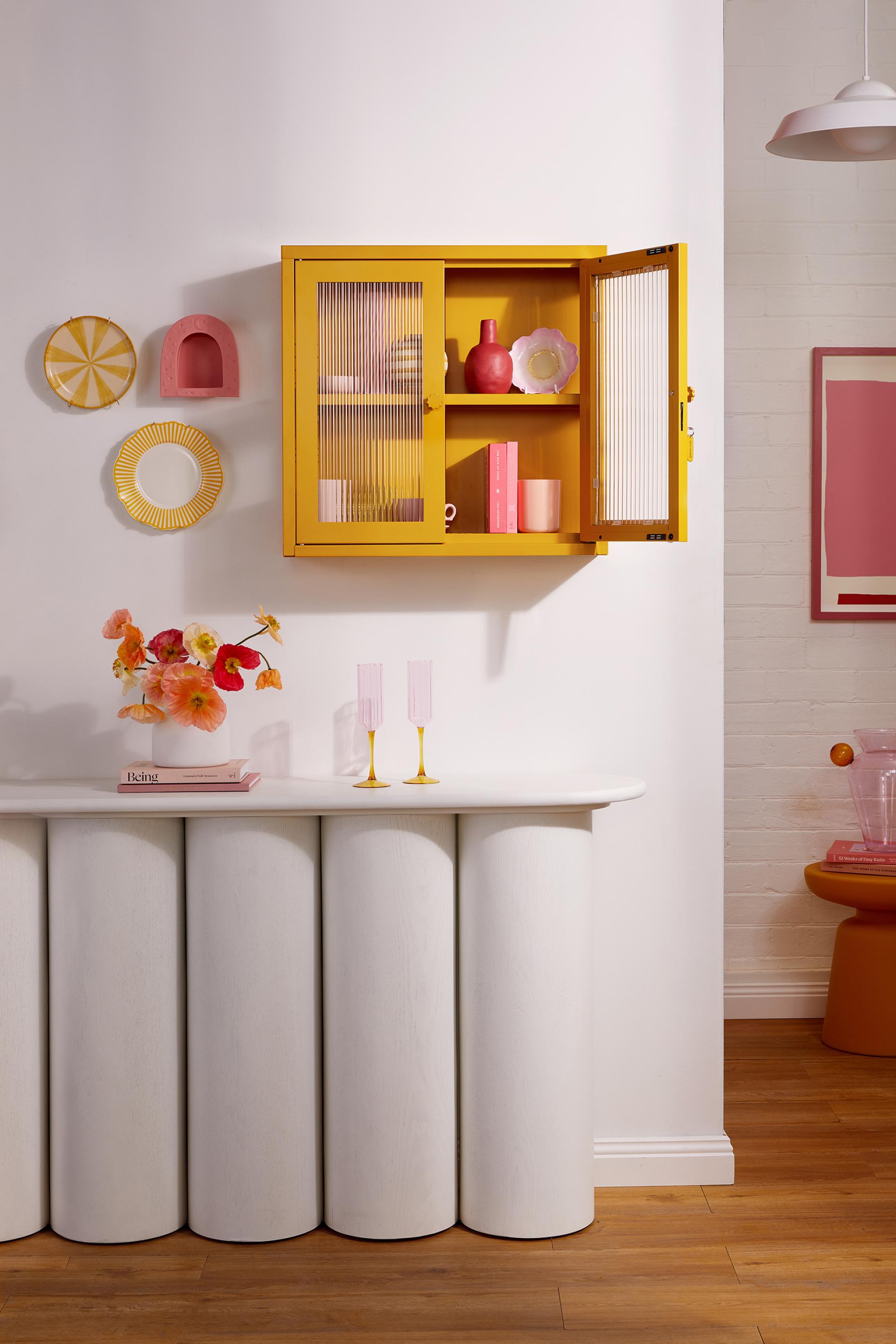 Colorful yellow cabinet from The Glass Collection with decorative items, flowers, and stylish tableware set against a bright wall.