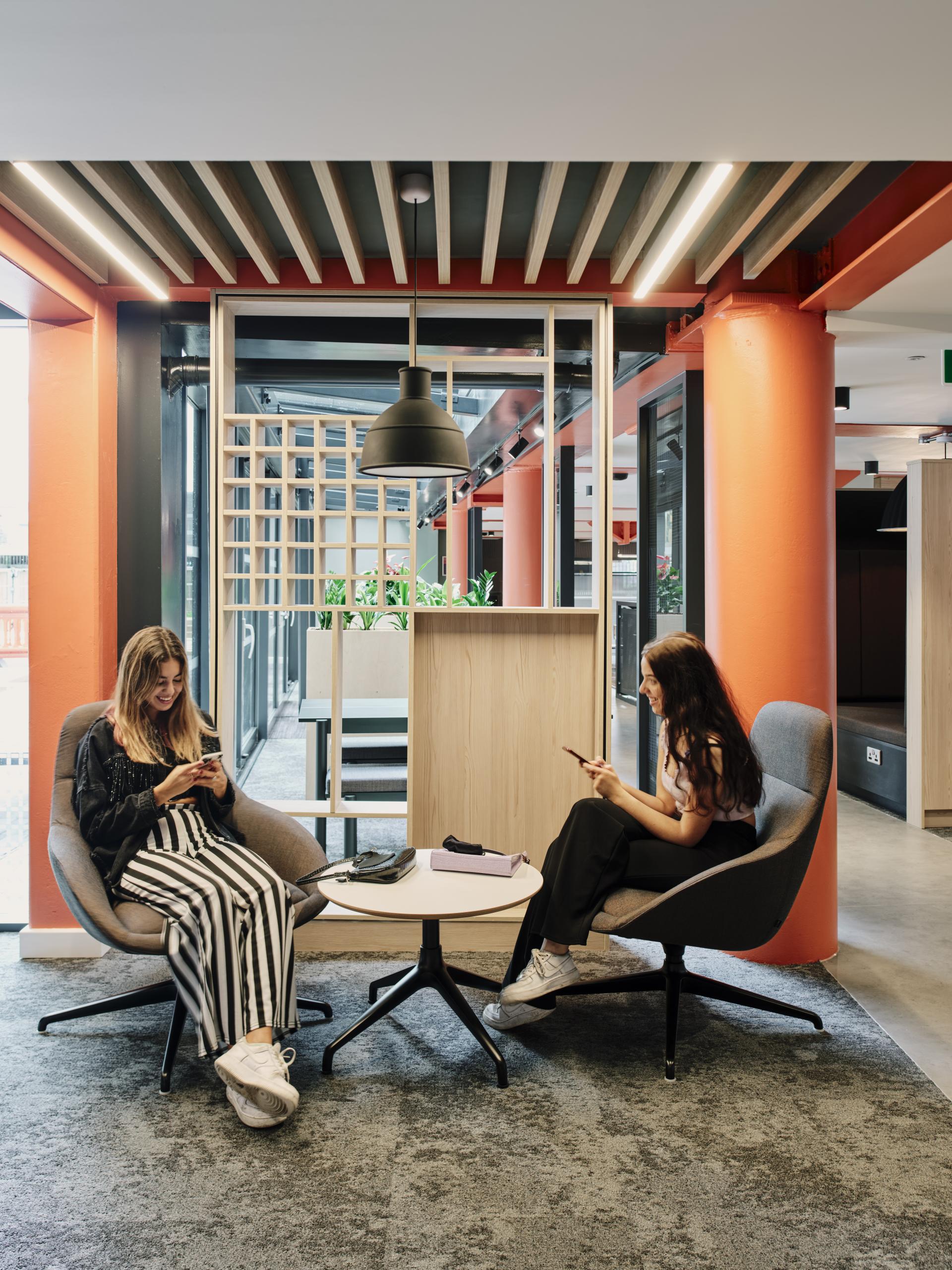 Two young women engaged in conversation on mobile devices in a modern, stylish workspace with vibrant decor.