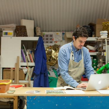 Ceramic artist Matthew Raw working at a cluttered studio desk, engaging with laptop and sketchbook amidst materials.