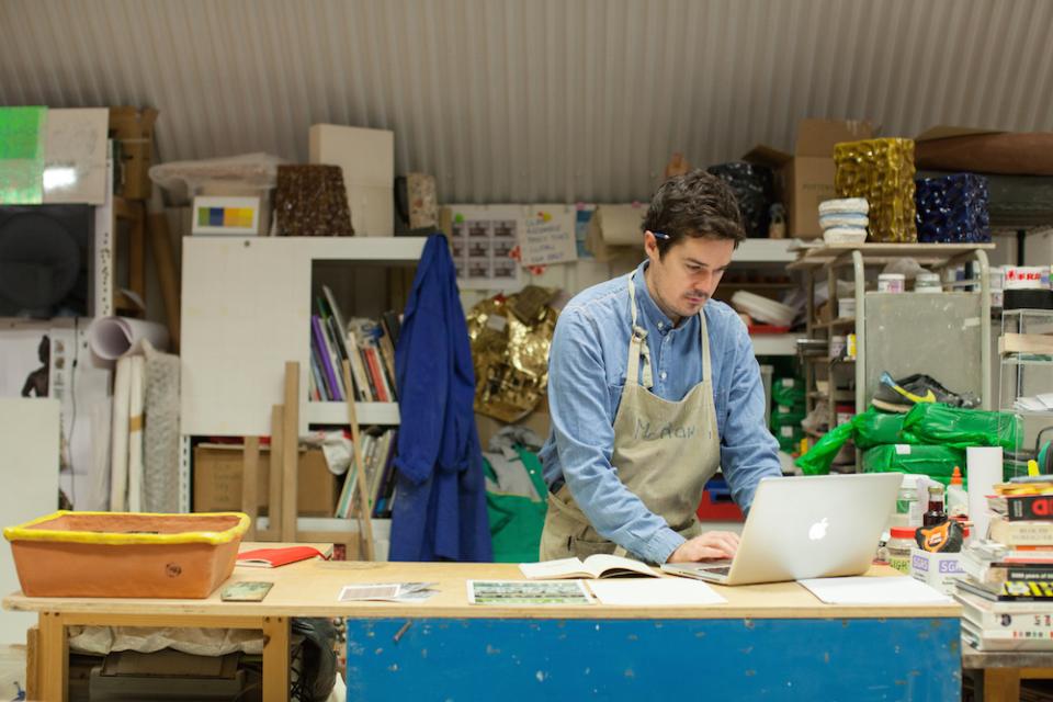 Ceramic artist Matthew Raw working at a cluttered studio desk, engaging with laptop and sketchbook amidst materials.