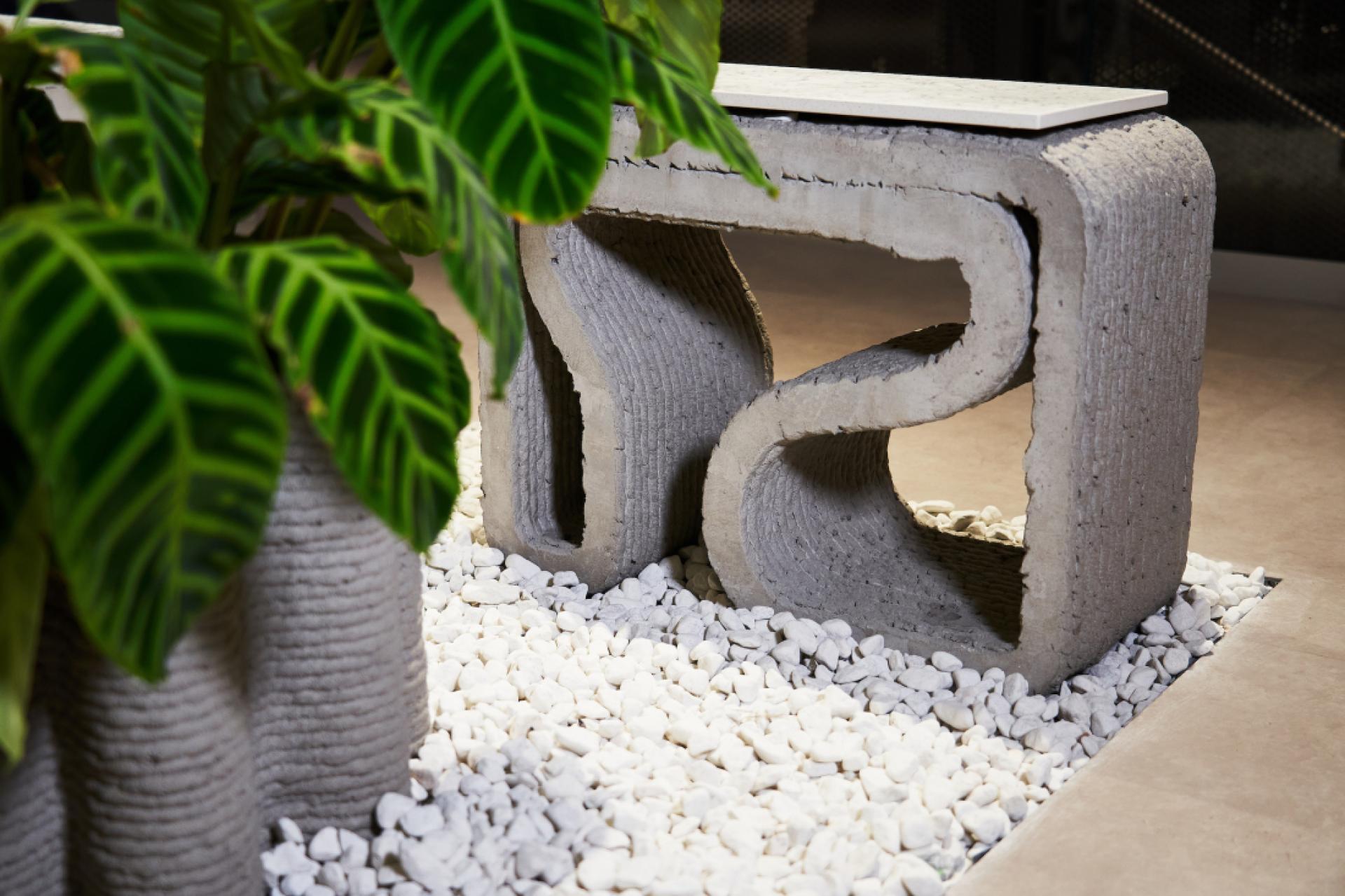 Innovative 3D-printed concrete furniture piece featuring a unique curved design, surrounded by decorative pebbles and greenery.