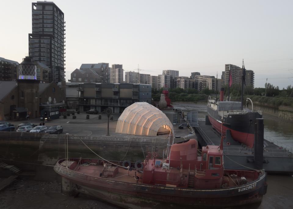 Innovative experimental CLT pavilion, The Armadillo, illuminated at dusk alongside historic boats and modern architecture.
