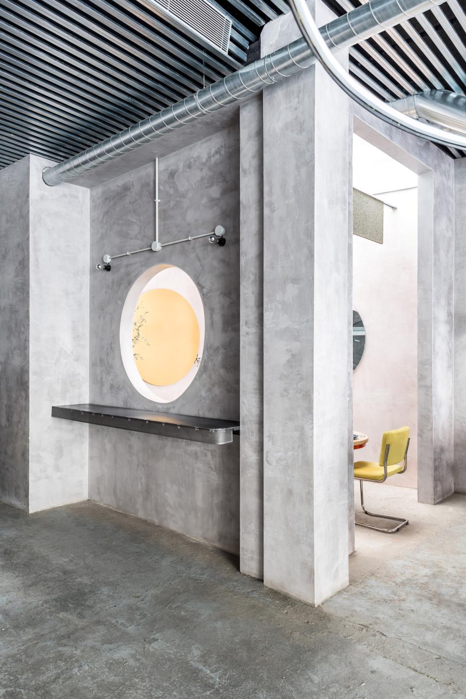 Contemporary restaurant interior featuring sleek concrete walls, a round window, and contrasting pastel furniture accents.
