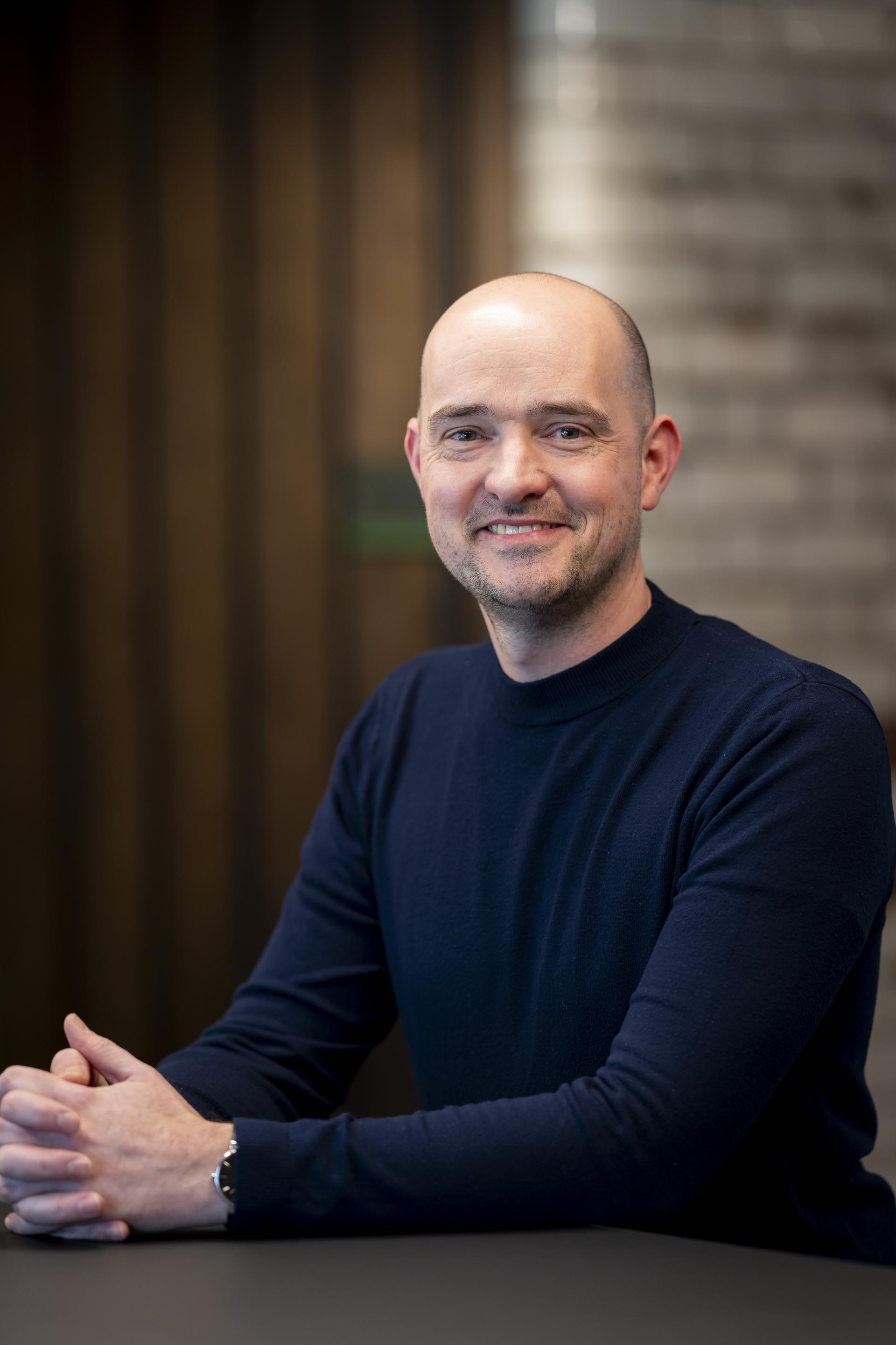 Smiling professional man in a navy sweater, sitting at a table, discussing technology's impact on sustainable design.