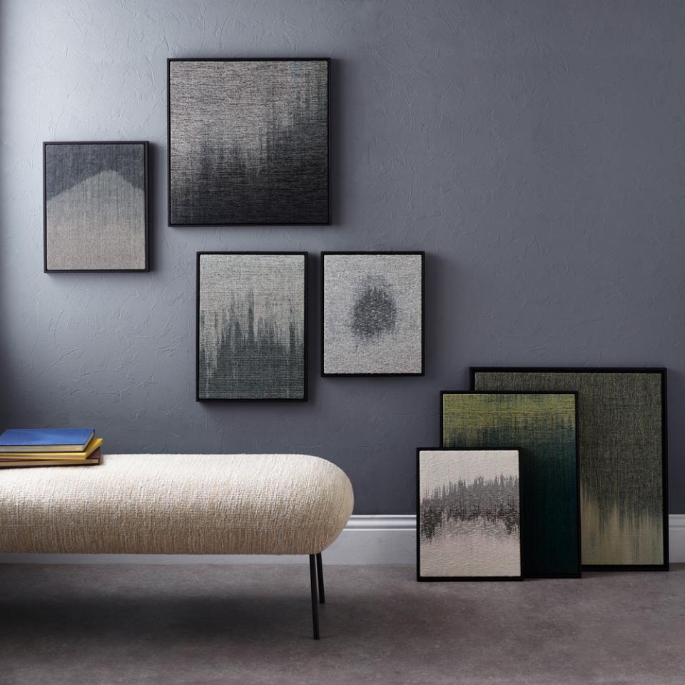 Framed textile artworks in various designs adorn a gray wall, accompanied by a textured bench and stacked books.