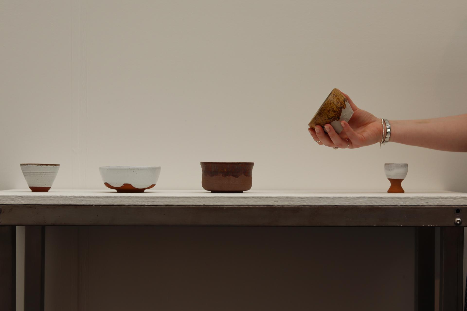 Hand reaching for a unique ceramic bowl among displayed pottery pieces at New Designers 2024 exhibition.