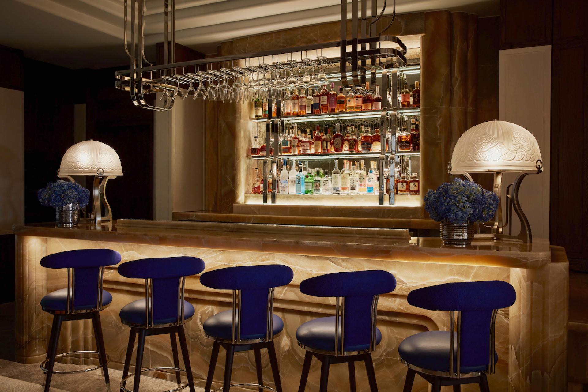 Elegant bar with blue seating, art deco elements, and a well-stocked liquor display at The Maybourne Bar.