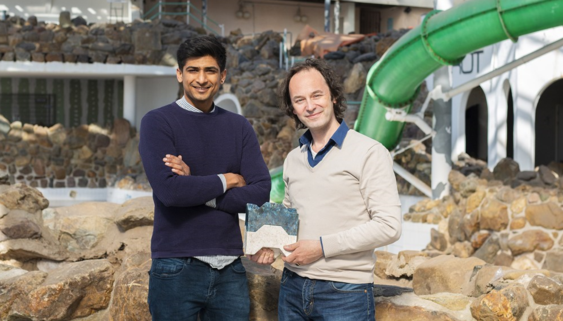 Two men proudly display a model of affordable housing made from recycled plastic, emphasizing sustainability and circular economy.