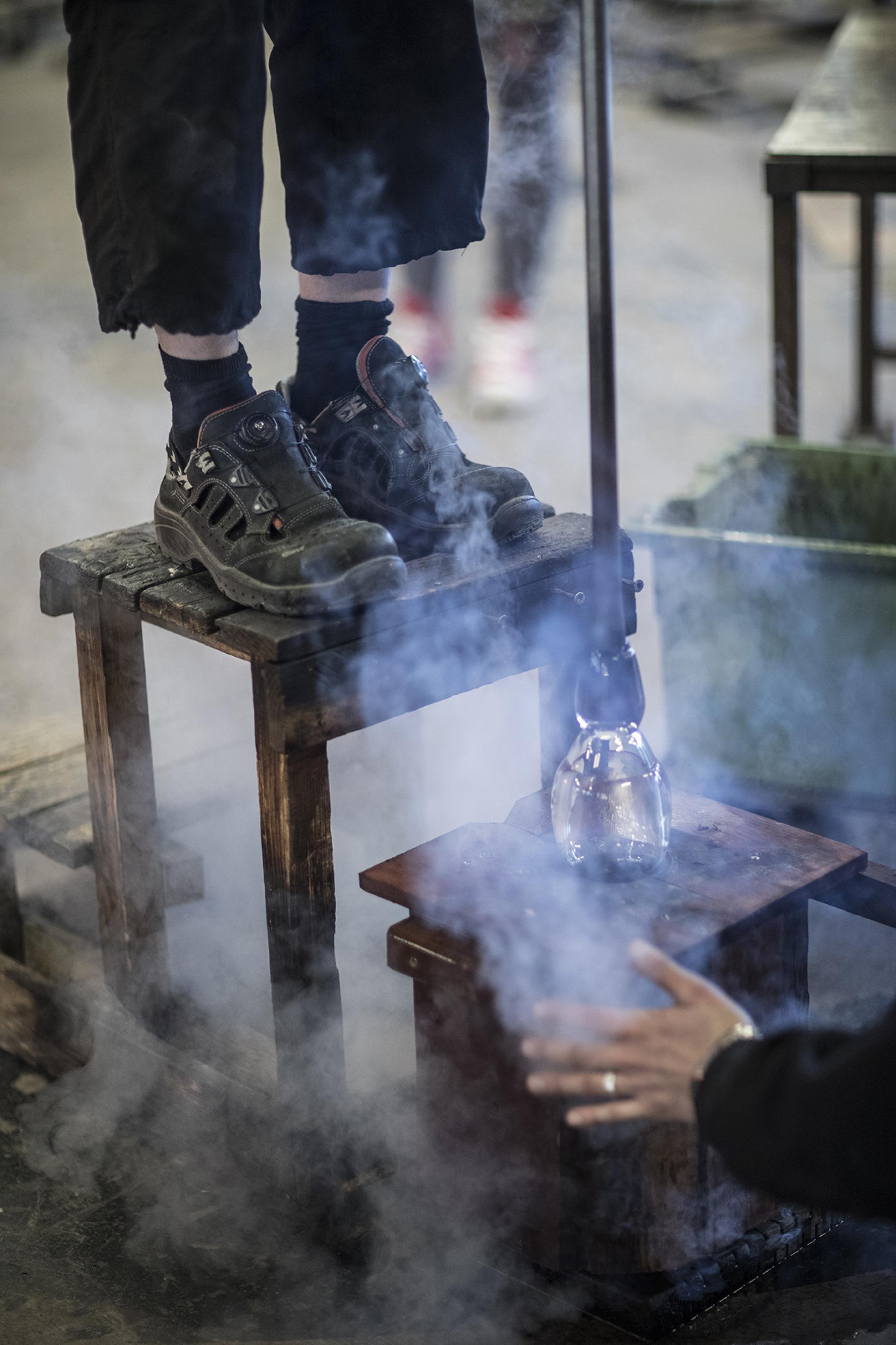 A glassblower stands on a wooden stool, shaping molten glass amid wisps of smoke at The Glass Factory in Småland.