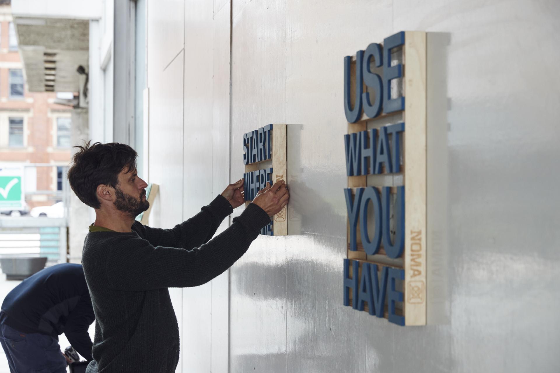 A man installs uplifting wooden signs with phrases in the NOMA neighborhood of Manchester, emphasizing community and creativity.