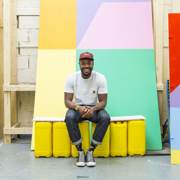 Yinka Ilori smiles while seated in a vibrant studio, surrounded by colorful furniture and geometric painted backgrounds.
