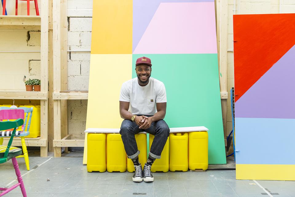 Yinka Ilori smiles while seated in a vibrant studio, surrounded by colorful furniture and geometric painted backgrounds.