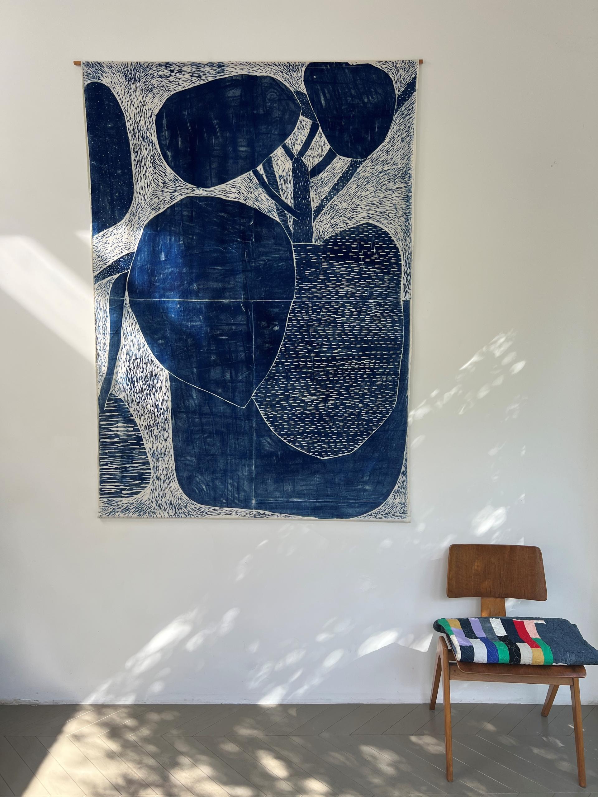 Textile wall art featuring abstract blue foliage design, displayed in an elegant gallery setting with a wooden chair.